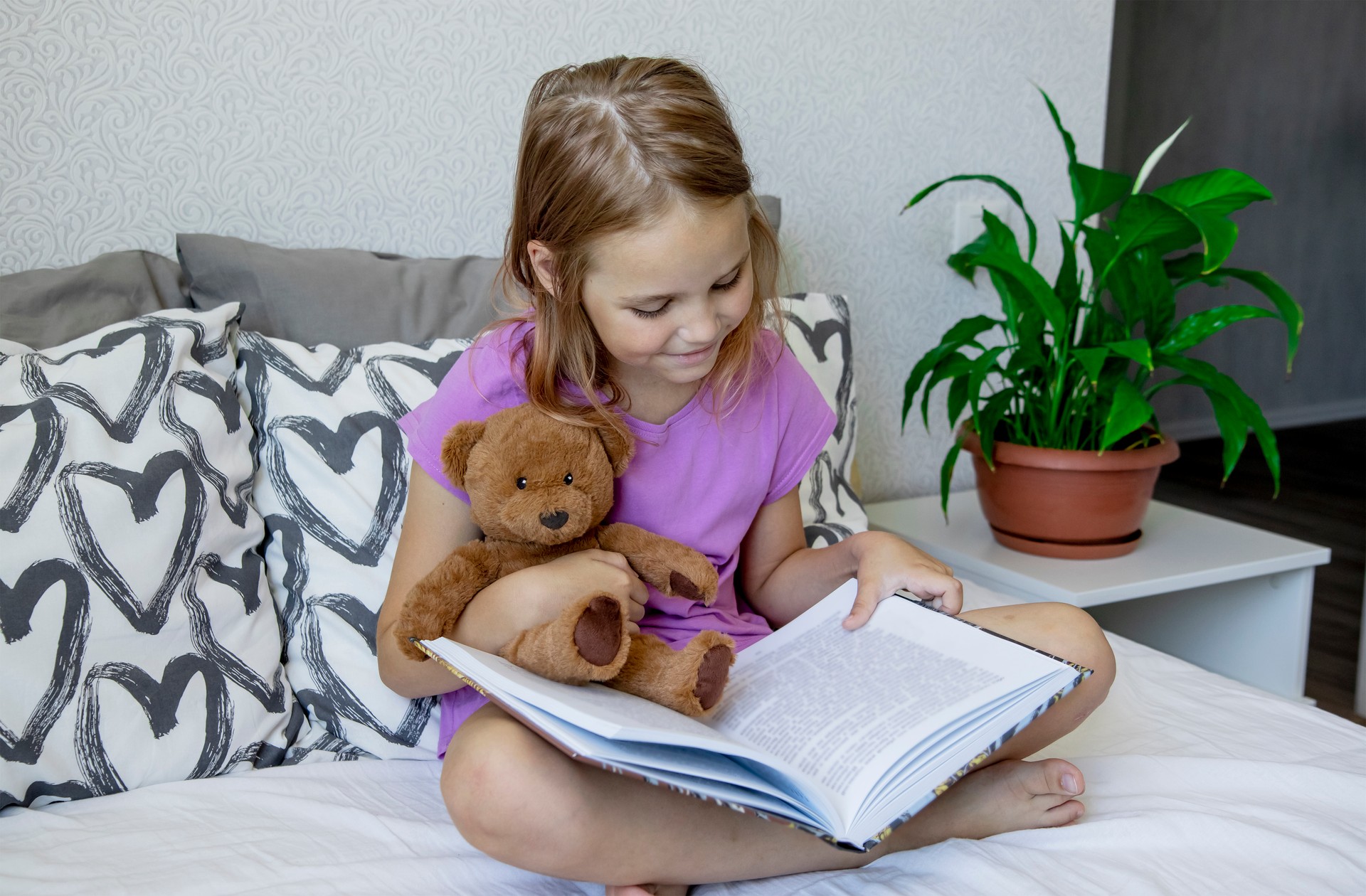 A little blonde girl is sitting on the bed before going to bed, hugging her teddy bear and reading him a children's fairy tale from a book.